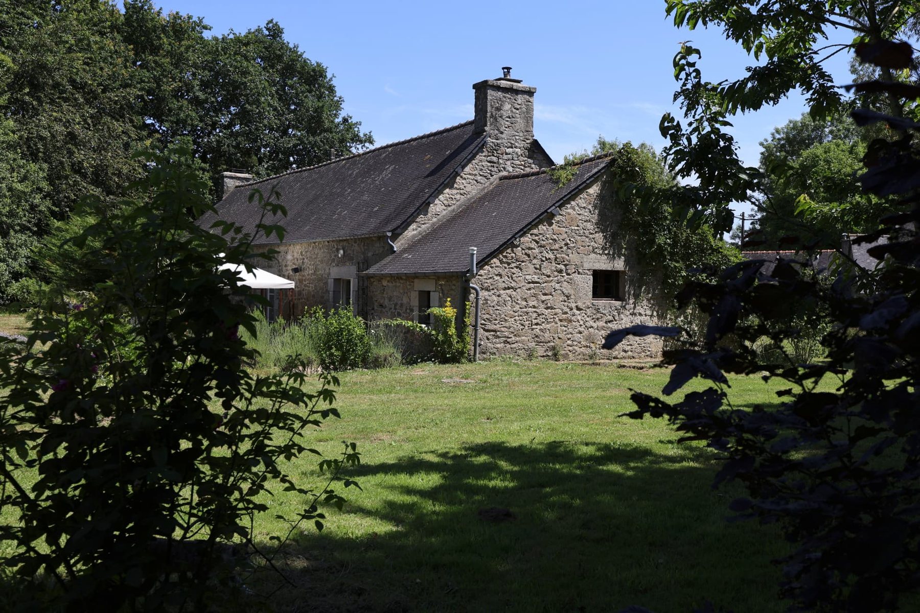 Stone house with slate roof seen across a sunlit lawn, framed by shaded foliage